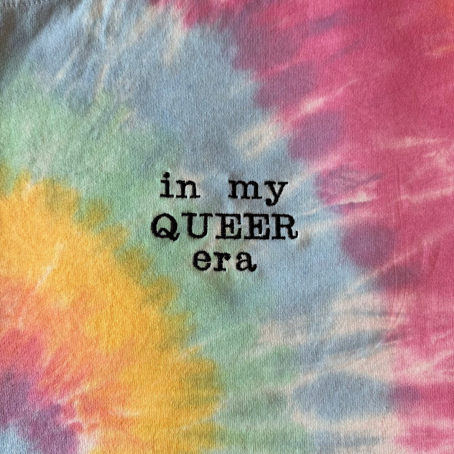 An upclose picture of a pastel tie dye shirt on. Off to the side of the shirt is the phrase "in my queer era" in black embroidery thread.