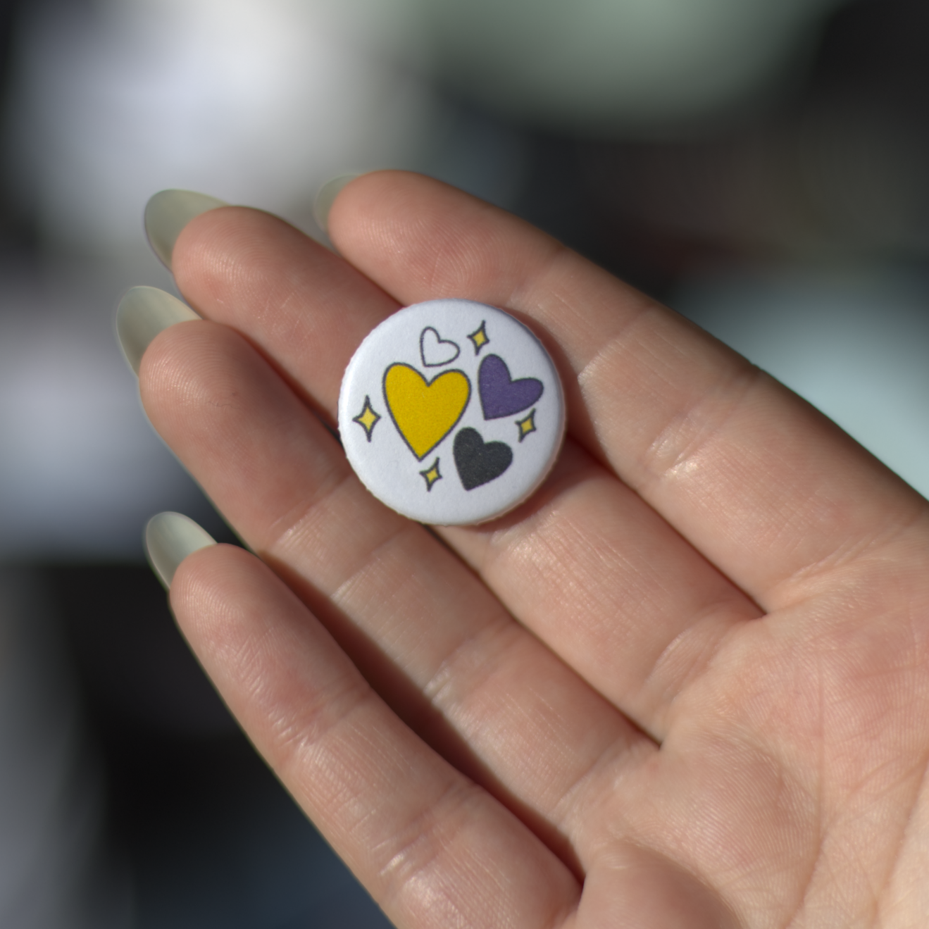the nonbinary hearts button, featuring four hearts in the nonbinary colors: white, yellow, purple, & black with four small sparkles around them.
