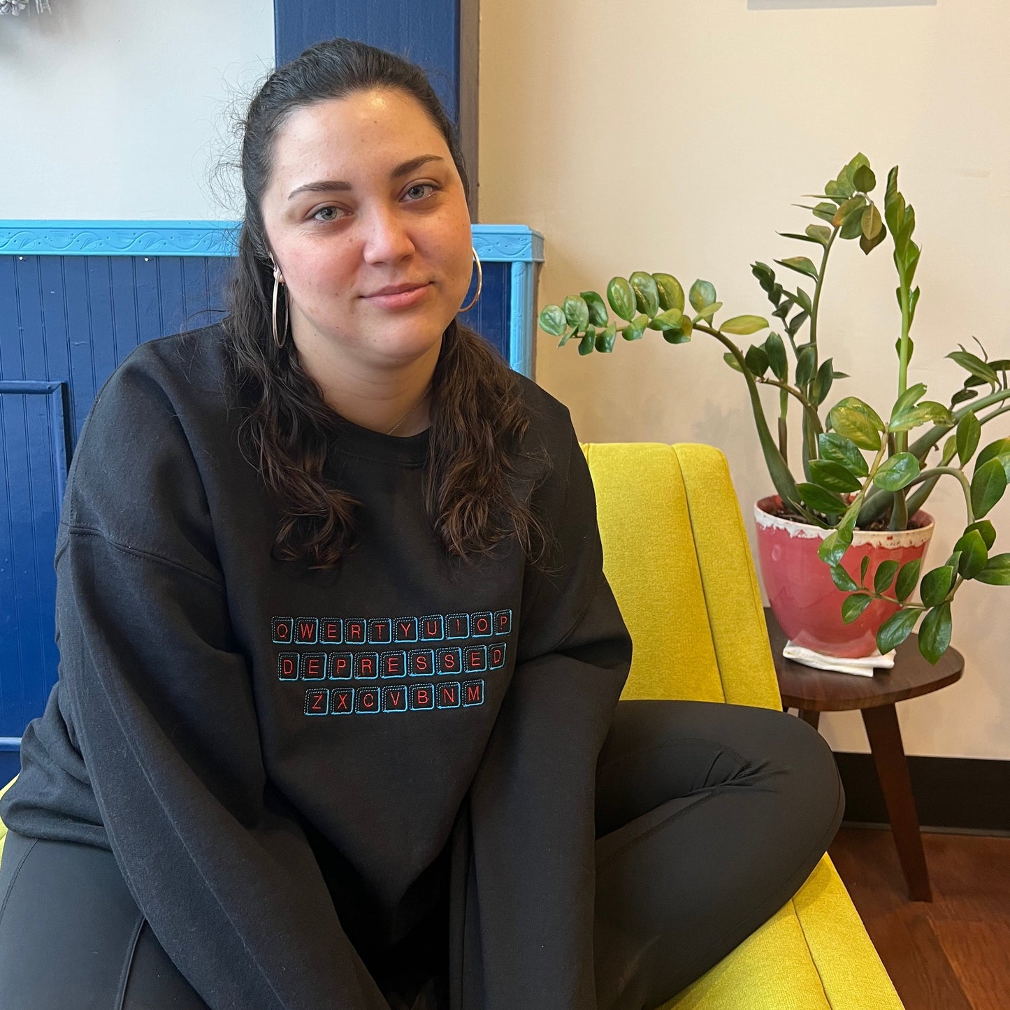 A girl with a gentle smile, sitting on a yellow chair next to a houseplant wearing a black crew neck. On the black crew neck is an embroidered design inspired by a qwerty keyboard but the middle row of keys are replaced with keys that spell DEPRESSED. The key caps are embroidered in a light blue thread, with the letters on the caps embroidered in a bright red thread.