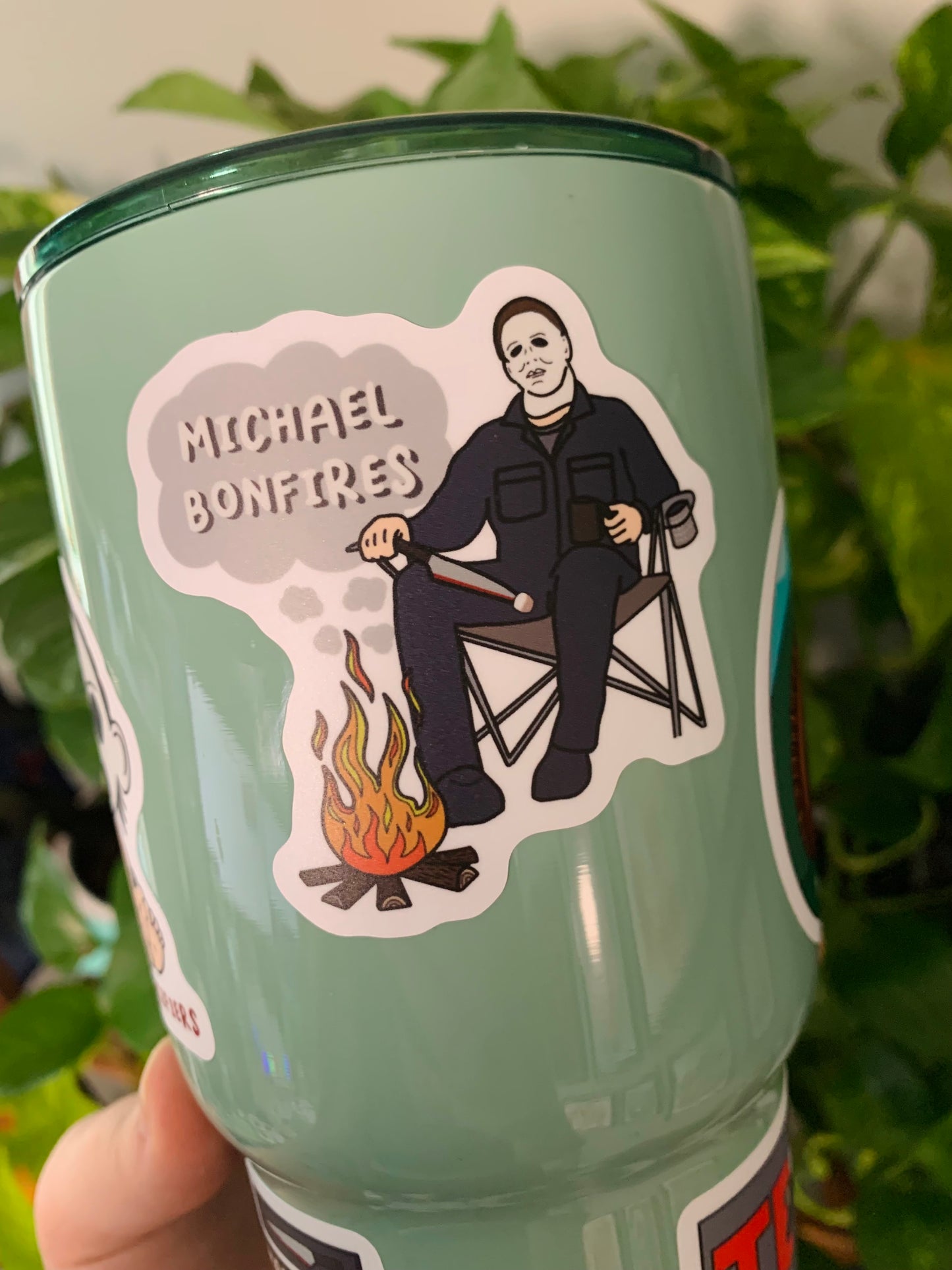 An up close picture of the Michael Bonfires sticker. The sticker features Michael Myers sitting in a camp chair, in front of a fire, holding a knife. On the tip of the knife there is a marshmellow for roasting over the fire. In smoke clouds above the fire is the words "Michael Bonfires".