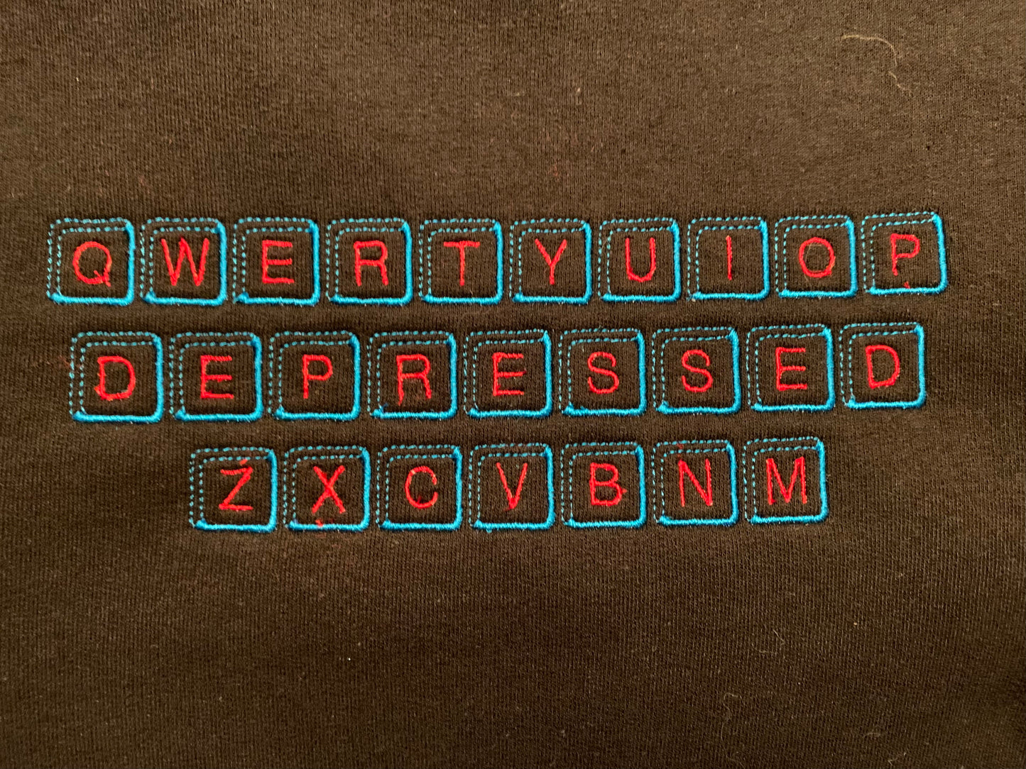 An upclose picture of a black crew neck with an embroidered design inspired by a qwerty keyboard on it. In the design, the middle row of keys are replaced with keys that spell DEPRESSED. The key caps are embroidered in a light blue thread, with the letters on the caps embroidered in a bright red thread.