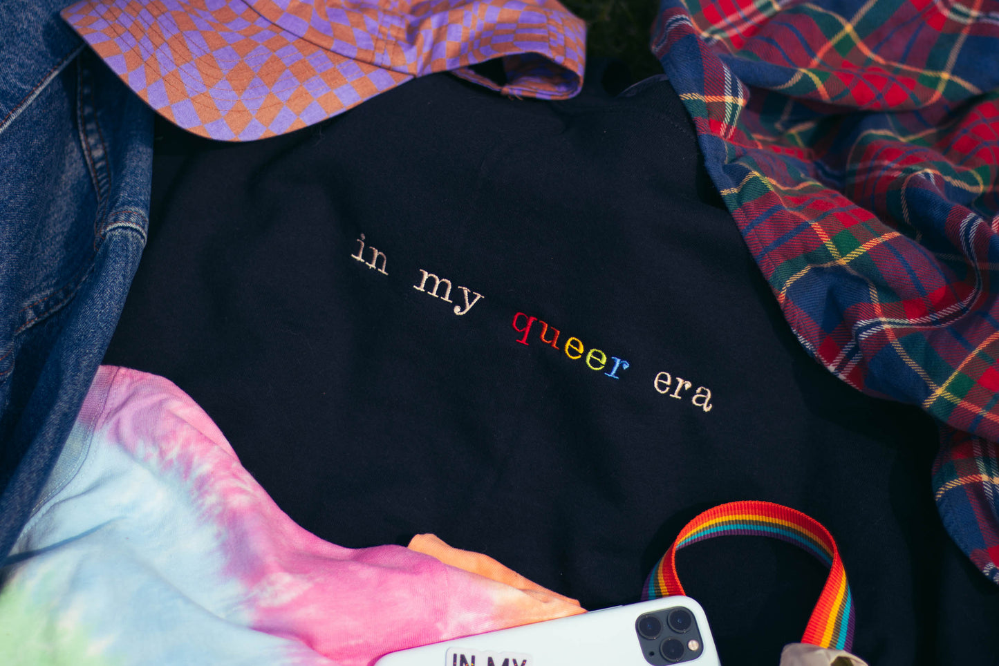 A closeup flatlay of various items, including a black crew neck with "in my queer era" embroidered on it. Surrounding the crew neck is various items, such as a checkered hat, a flannel, a phone, a jean jacket, & a tie dye shirt.