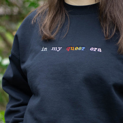 A closeup picture of a torso, wearing a black crew neck. On the crew neck is the words "in my queer era" embroidered in cream and multicolored thread.