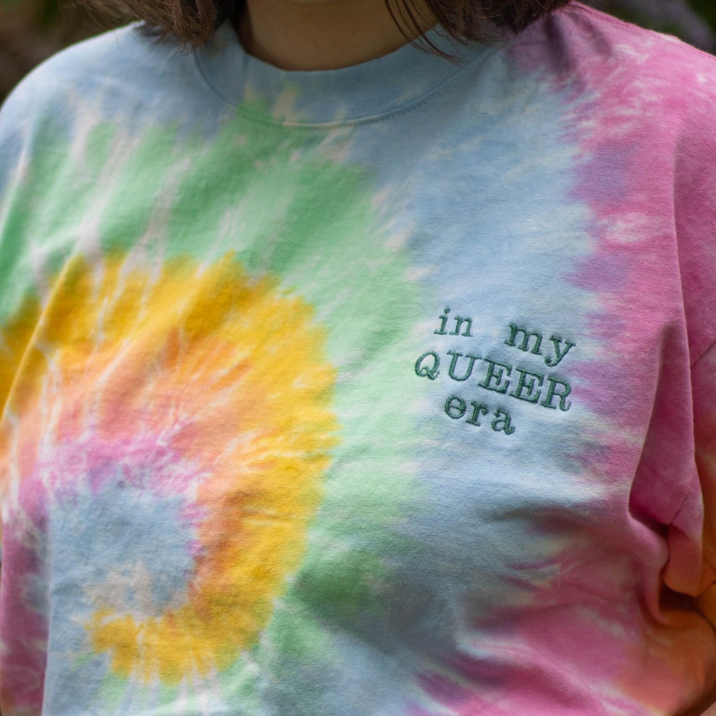 An upclose picture of a torso with a pastel tie dye shirt on. Off to the side of the shirt is the phrase "in my queer era" in green embroidery thread.