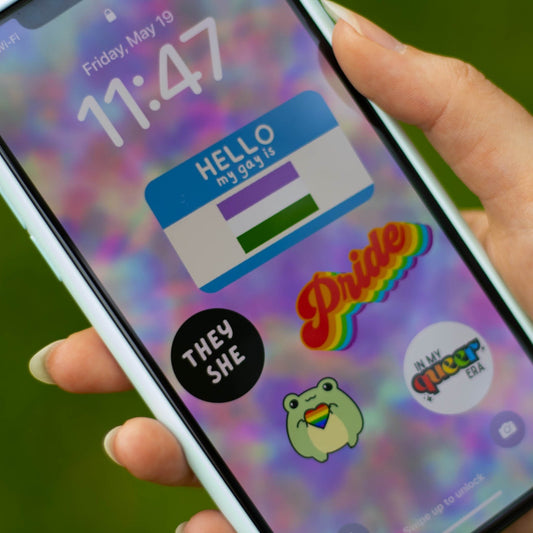 A picture of a phone with the HELLO my gay is wallpaper. The wallpaper is a blurry tie dye background with a name tag design that says "Hello my name is" written above and a pride flag featured in the name section. There is also a black circle with selected pronouns on it. To the right of that is a colorful design of the word Pride. Underneath is a small cartoon frog holding a rainbow filled heart. Lastly, at the bottom right is a white circle with the in my queer era within it. 
