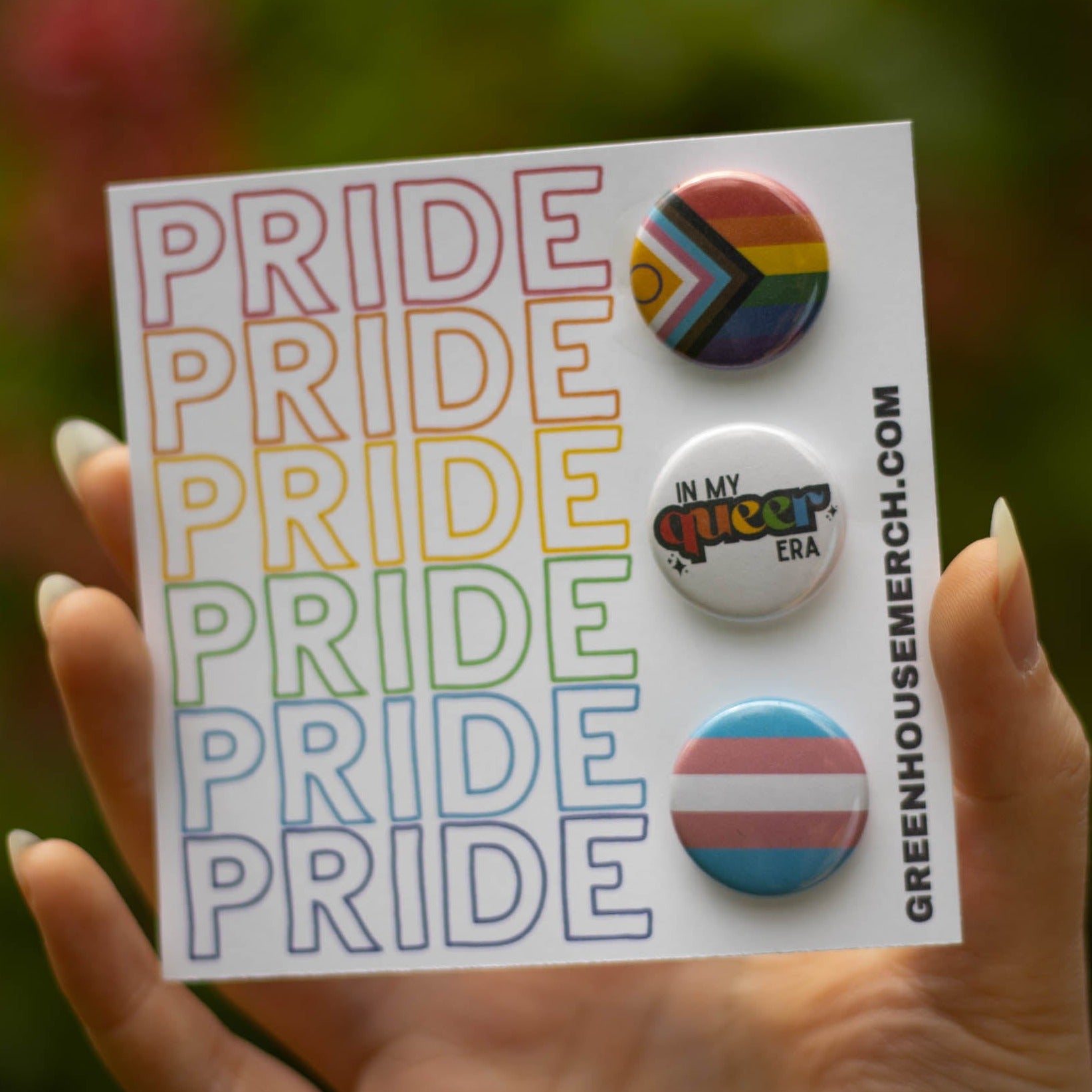 Up close picture of the Pride Button Pack. The backing card is a white cardstock with the word PRIDE multiple times down the left side. On the card features the inclusive progress flag pin, the in my queer era pin, and the transflag pin.