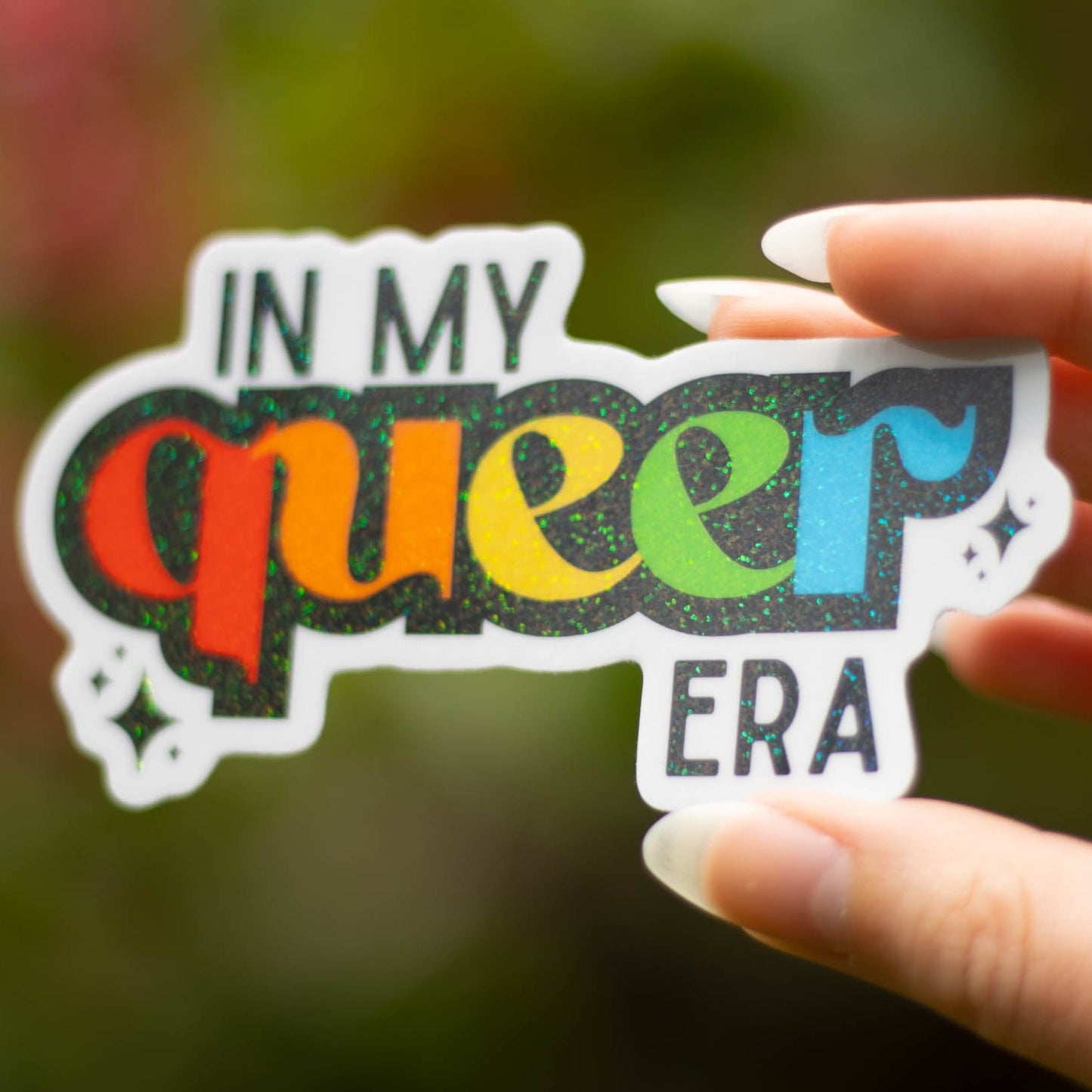 a close up picture of the in my queer era sticker. the sticker is the phrase in my queer era. the word queer is in a font with small flourishes around the edges. Each letter of the word queer is a different color from red, orange, yellow, green and blue. Around the word are two sets of sparkles.