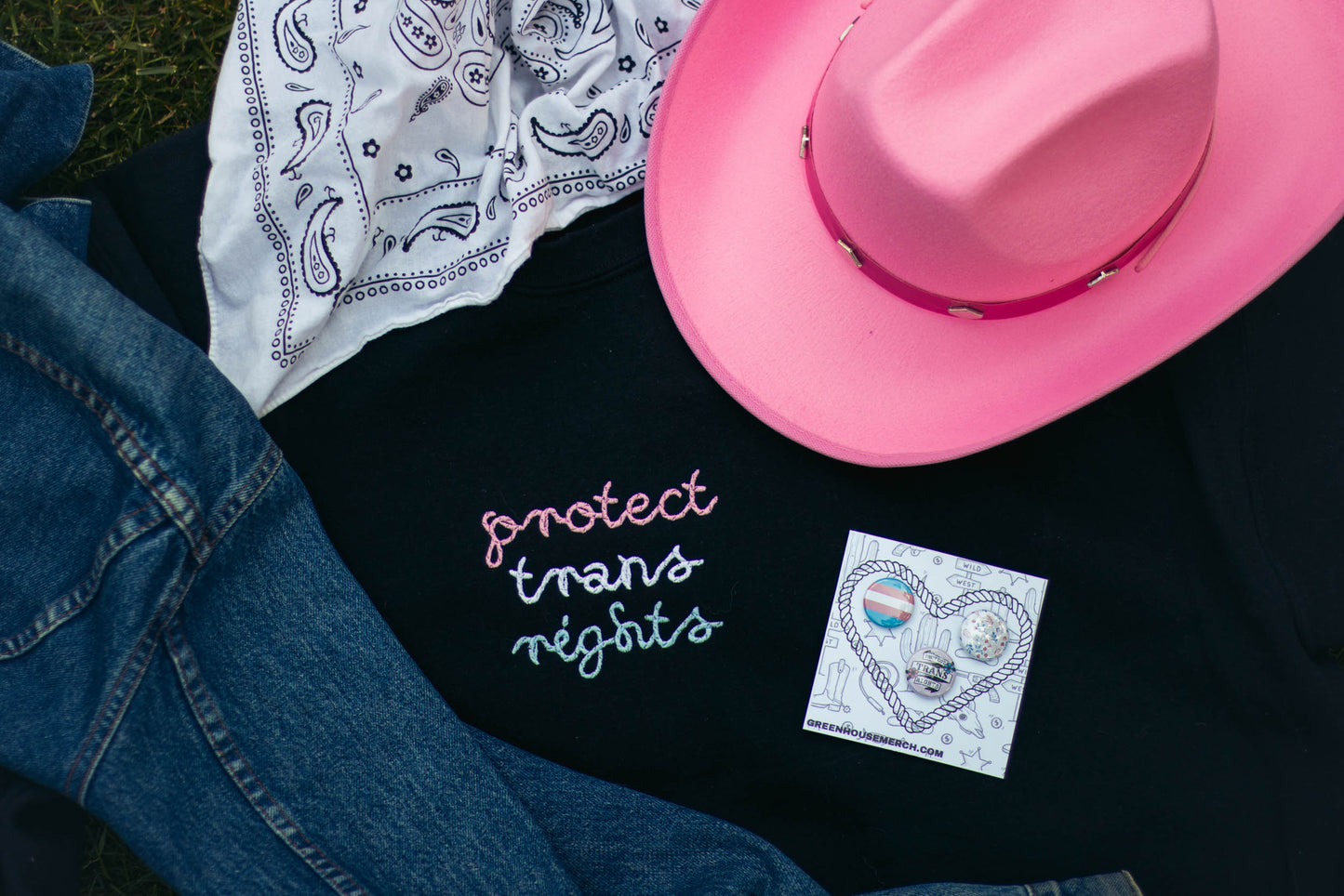 A flatlay image of the protect trans rights crew neck surrounded by various items. The protect trans rights crew neck is a black crew neck with the phrase "protect trans rights" in a rope style font in the trans flag colors. Surrounding the crew is a medium wash jean jacket, a white bandana, a bright pink cowboy hat, and the protect trans rights button pack.