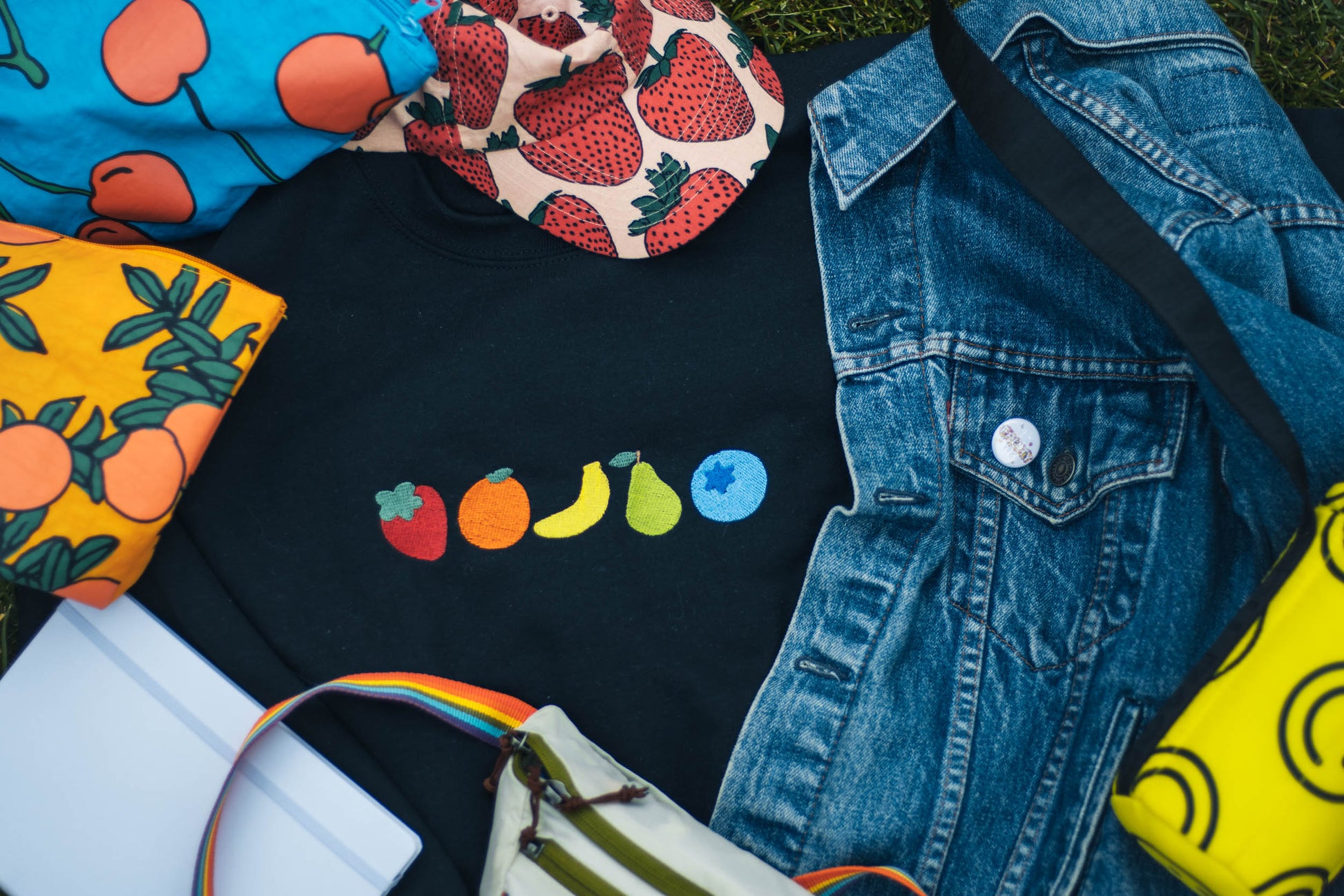 A flatlay picture of the fruity crewneck surrounded by many colorful and fruity items. The fruity crewneck features 5 very simple embroidered fruits. From left to right the fruits are strawberry, orange, banana, pear, and a blueberry. Surrounding the fruity crew neck is a strawberry hat, a jean jacket with a pin, a smiley face bag, a fanny pack with a rainbow strap, a lavender journal, and two fruit themed baggies.