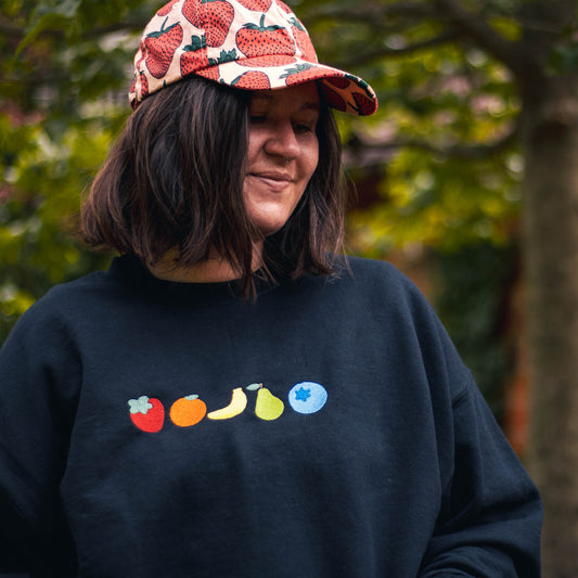 A picture of Bella from the waist up looking gently to the side while wearing a strawberry baseball cap. She is wearing the fruity crew neck which features 5 very simple embroidered fruits. From left to right the fruits are strawberry, orange, banana, pear, and a blueberry.