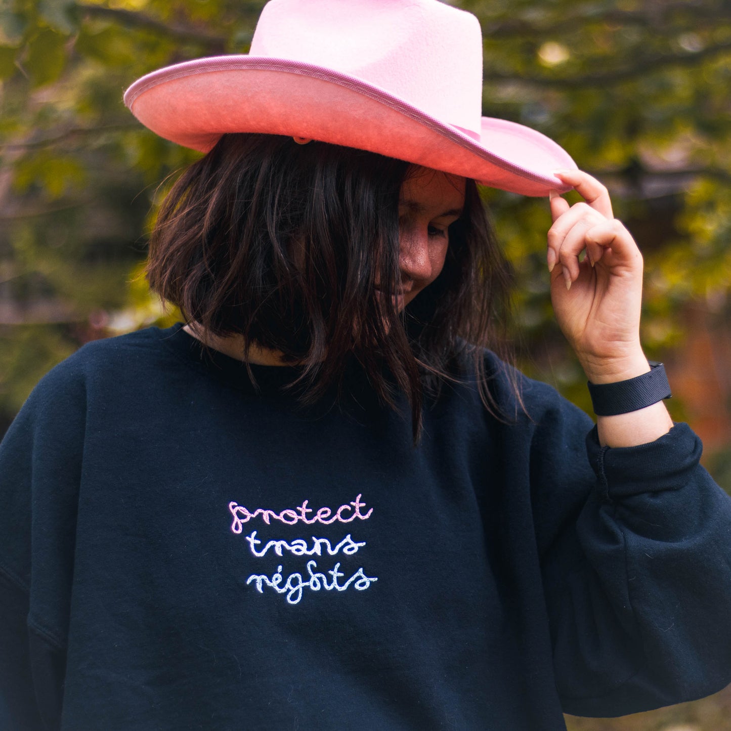 Bella is looking to the side tipping a bright pink cowboy hat and gently smiling. They are wearing a black crew neck with "protect trans rights" in a rope style font in trans flag colors.