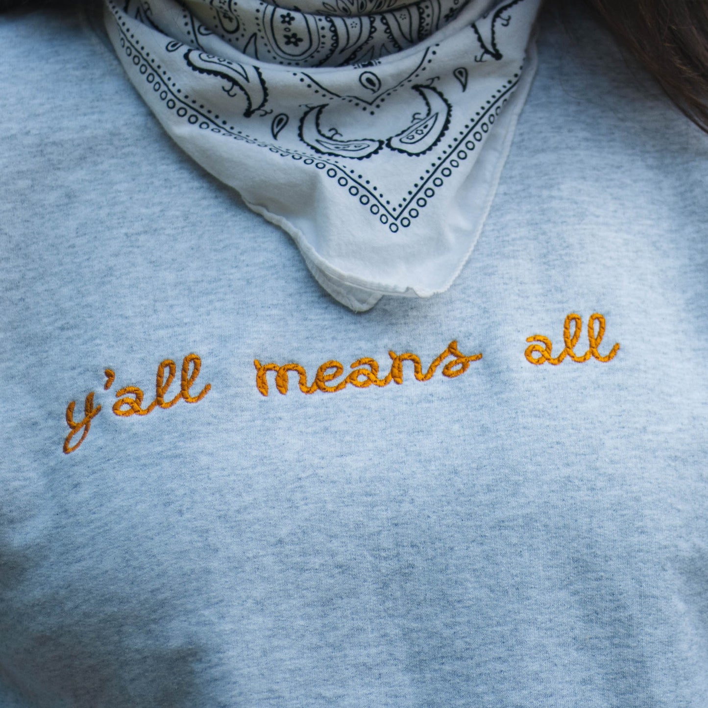 A close up picture of Ariel wearing the "y'all means all" crewneck. The crewneck is a light grey color with the phrase y'all means all in a rope style font in a rich yellow color.