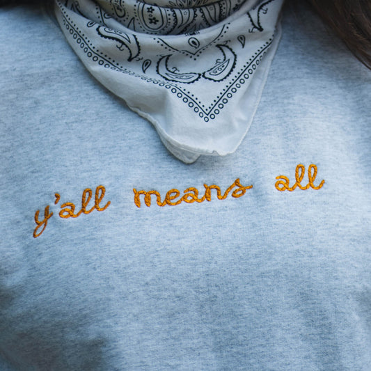 A close up picture of Ariel wearing the "y'all means all" crewneck. The crewneck is a light grey color with the phrase y'all means all in a rope style font in a rich yellow color.