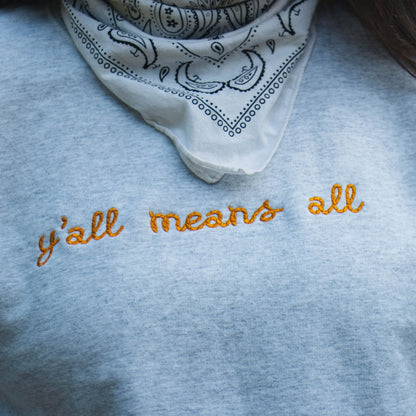 A close up picture of Ariel wearing the "y'all means all" crewneck. The crewneck is a light grey color with the phrase y'all means all in a rope style font in a rich yellow color.