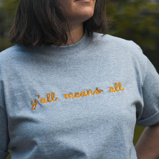 A close up picture of Bella wearing the "y'all means all" tshirt. The tshirt is a medium grey color with the phrase y'all means all in a rope style font in a rich yellow color.