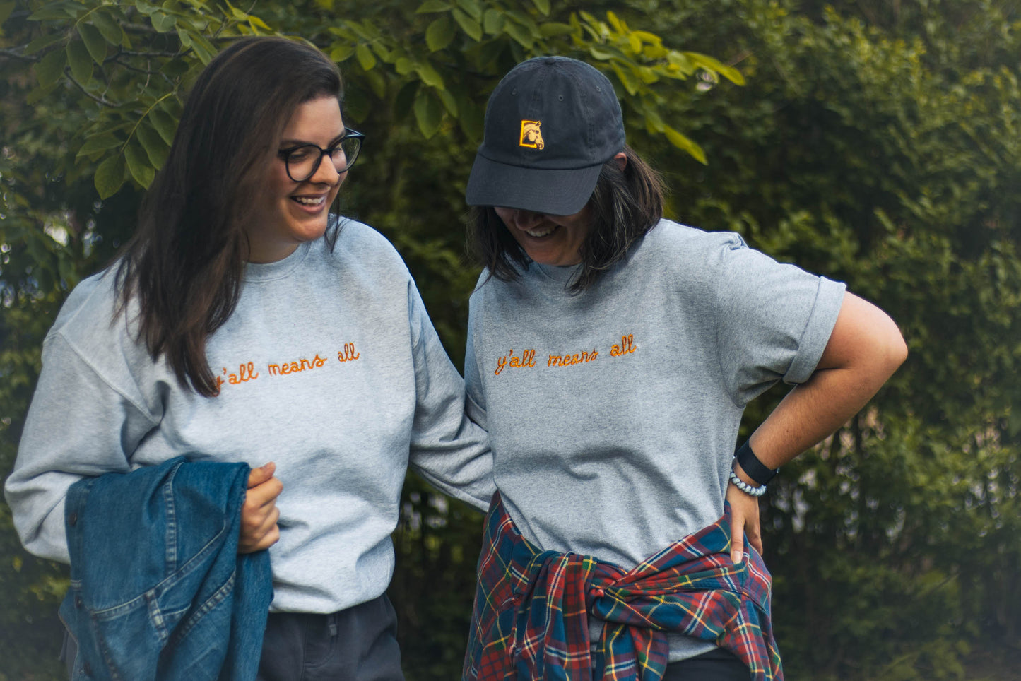 Ariel and Bella standing side by side laughing. Both are wearing the "y'all means all" designs. Ariel is wearing a crewneck in the size Large, and Bella is wearing the tshirt in the size Large.