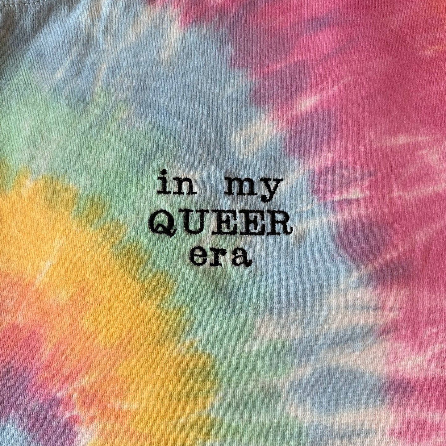 An upclose picture of a pastel tie dye shirt on. Off to the side of the shirt is the phrase "in my queer era" in black embroidery thread.