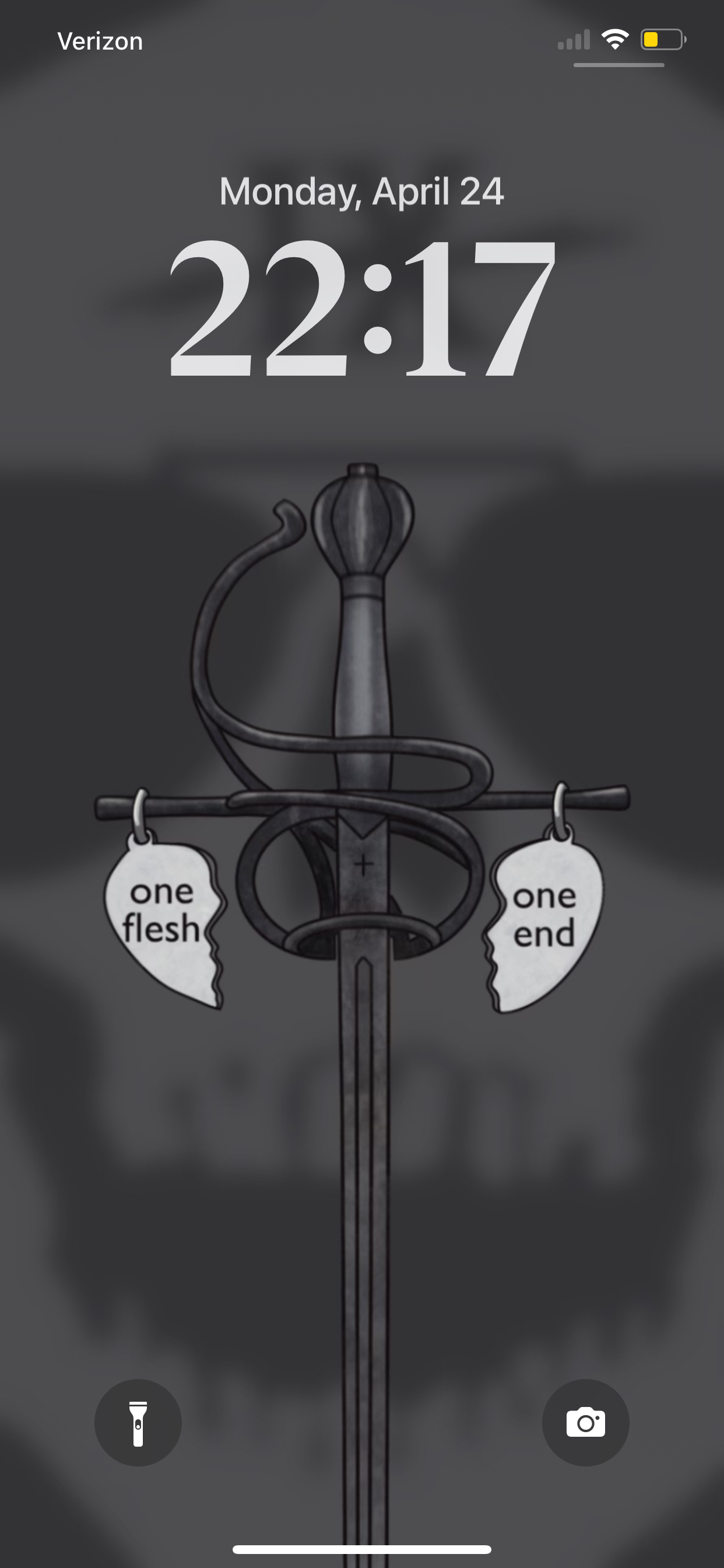 Image of phone lockscreen showing the "one flesh one end" wallpaper. The wallpaper is a black and grey design showcasing the handle of a black metal rapier with two charms hanging on each side of the handle. Each charm is in the shape of a broken heart and has one flesh on the left heart, and one end on the right.