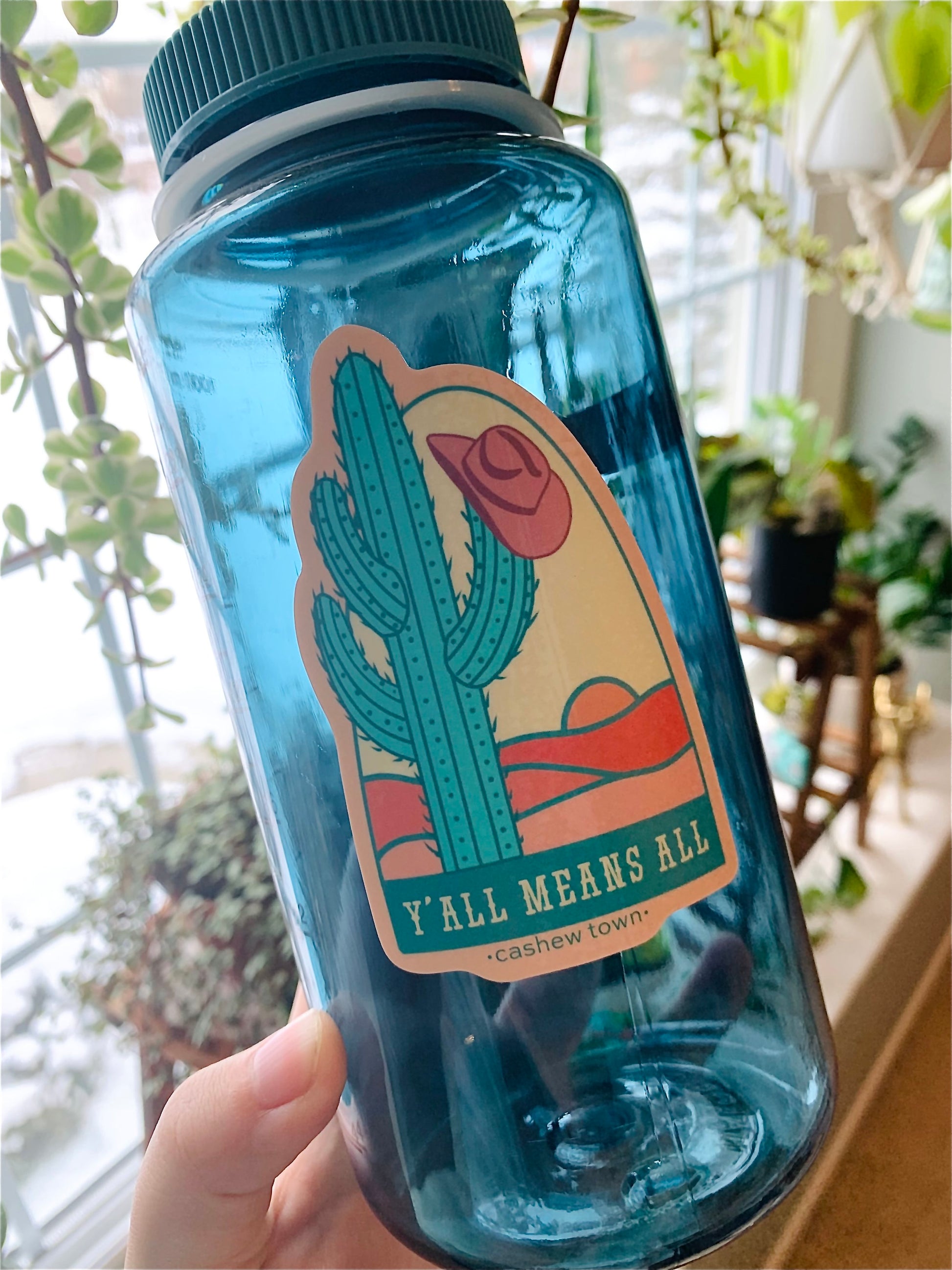 Our Y'all Means All sticker featured on a blue nalgene water bottle taken in front of houseplants on a window sill.