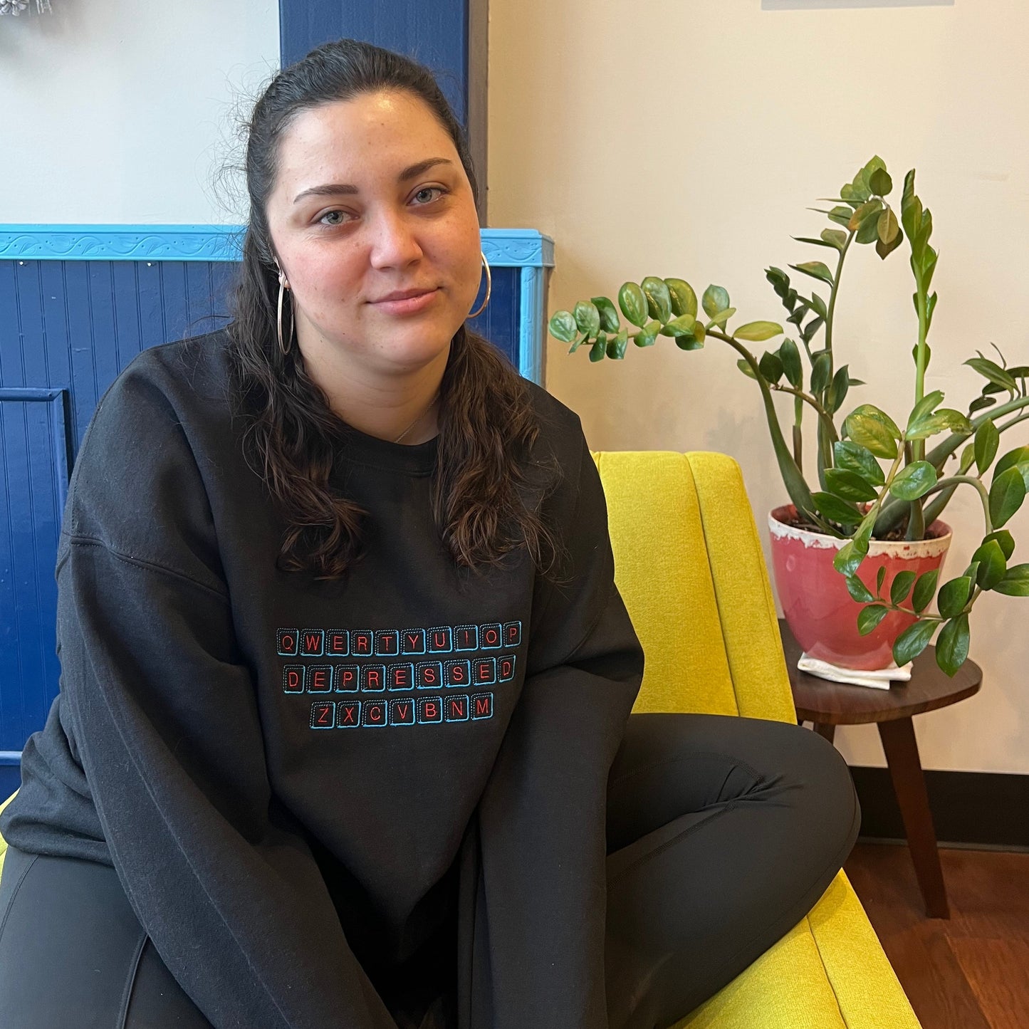 A girl with a gentle smile, sitting on a yellow chair next to a houseplant wearing a black crew neck. On the black crew neck is an embroidered design inspired by a qwerty keyboard but the middle row of keys are replaced with keys that spell DEPRESSED.  The key caps are embroidered in a light blue thread, with the letters on the caps embroidered in a bright red thread.