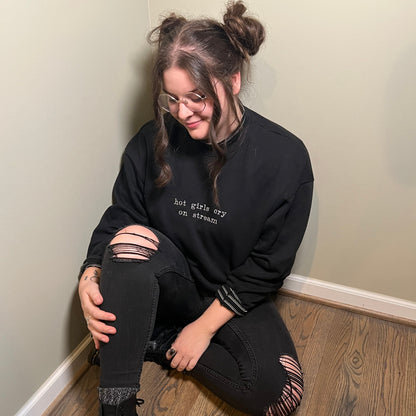 Ariel is sitting on the floor wearing a black crew neck with "hot girls cry on stream" embroidered on the front.