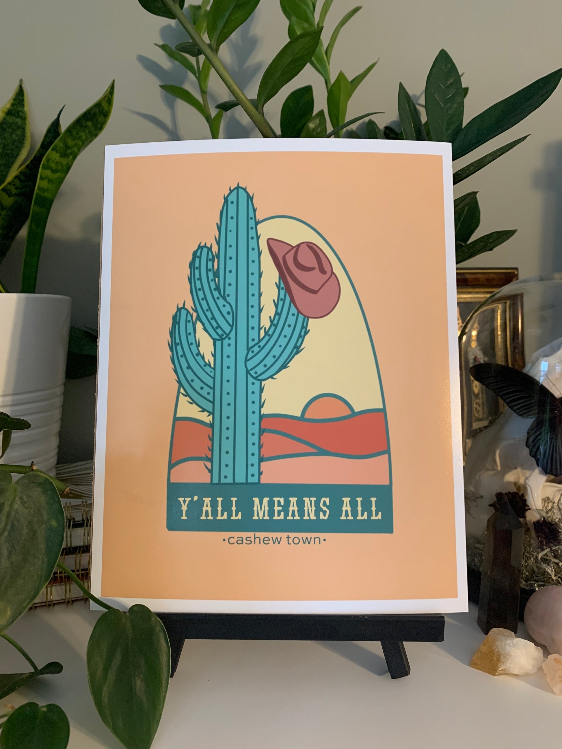 A print of the Y'all Means All design in front of house plants. The Y'all Means All Design is logo on a muted pastel orange background. The logo is a drawn green cactus with a muted maroon cowboy hat hanging off the side, in front of a very simple line color fill of a landscape with "Y'ALL MEANS ALL" underneath & "cashew town" in small letters under the design.