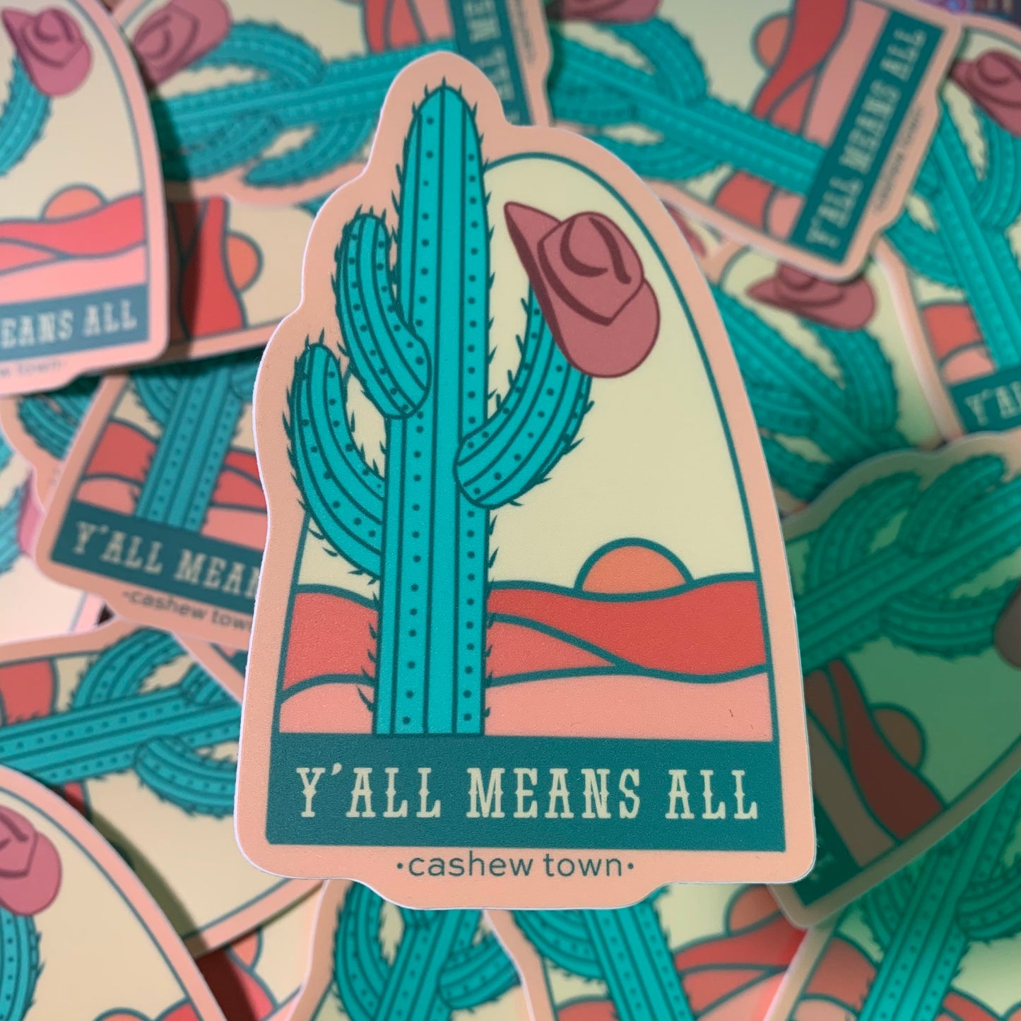 A sticker of a drawn green cactus with a muted maroon cowboy hat hanging off the side, in front of a very simple line color fill of a landscape with "Y'ALL MEANS ALL" underneath & "cashew town" in small letters under the design.
