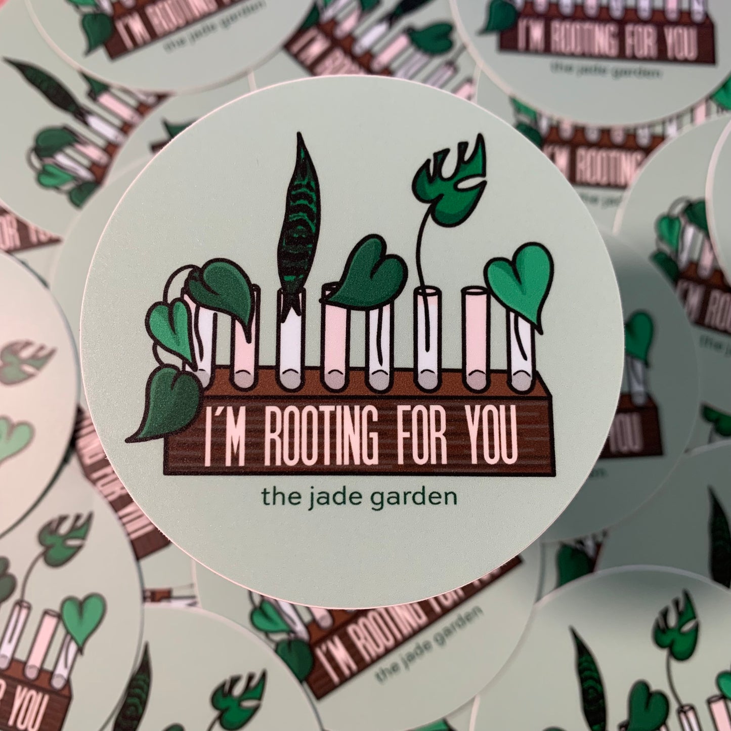 A sage green sticker, with a drawn design of a propagation station with leaves within the glass vials. "I'M ROOTING FOR YOU" is written on the wooden stand of the propogation station with "the jade garden" featured underneath.