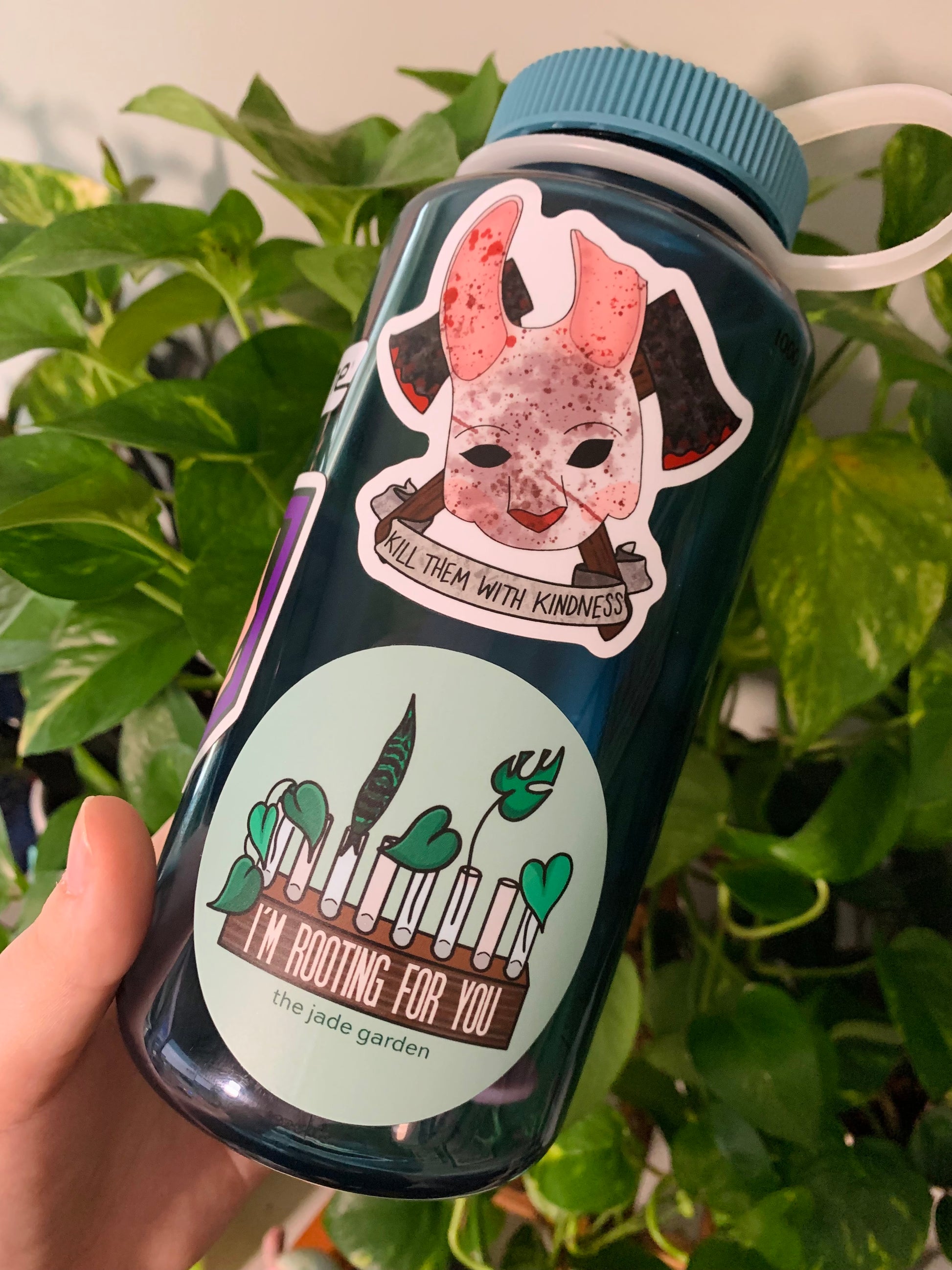 A blue nalgene water bottle in front of plants, with the Kill Them With Kindness Huntress Sticker & I'm Rooting For You propagation sticker on the side.