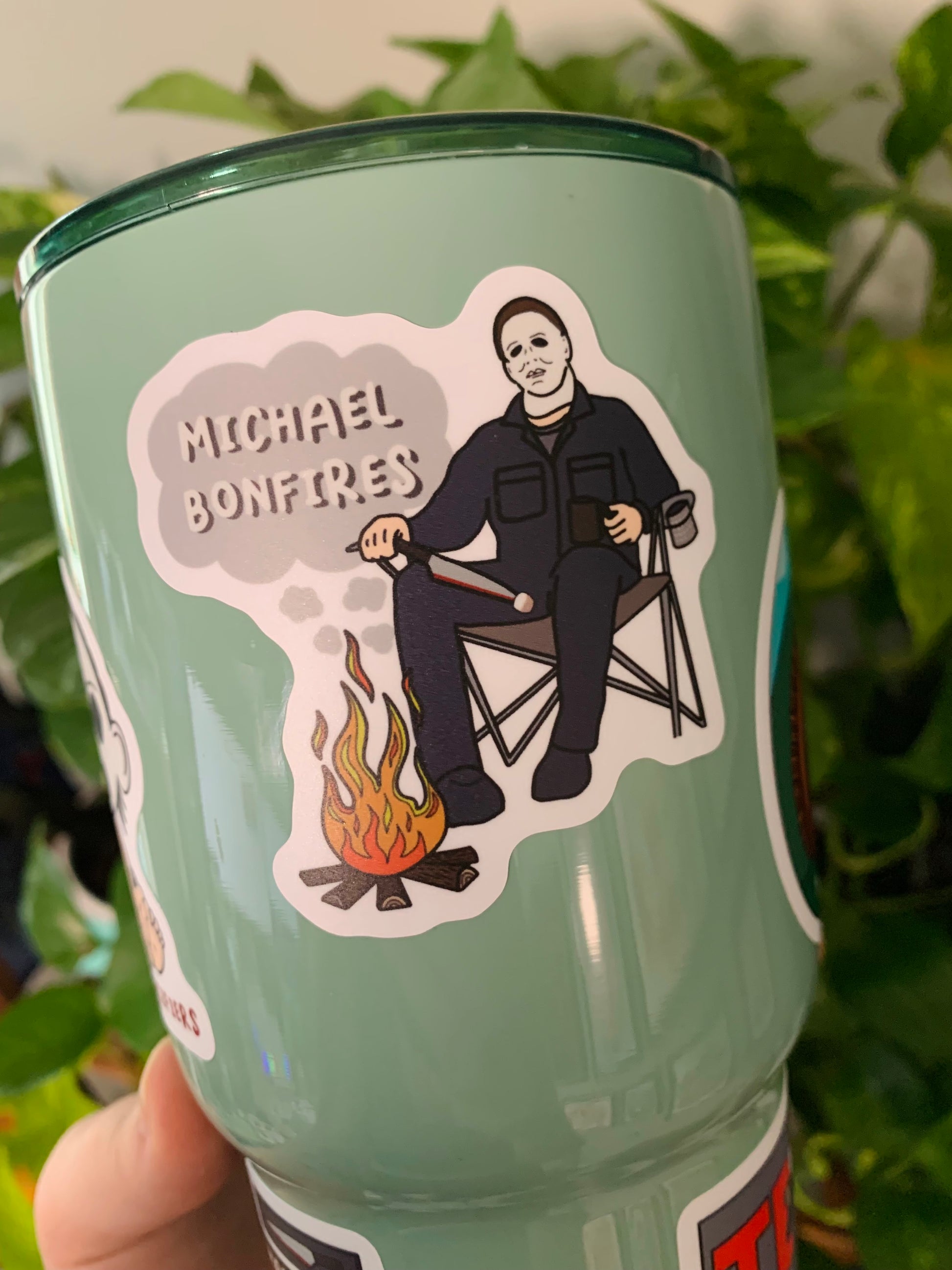 An up close picture of the Michael Bonfires sticker. The sticker features Michael Myers sitting in a camp chair, in front of a fire, holding a knife. On the tip of the knife there is a marshmellow for roasting over the fire. In smoke clouds above the fire is the words "Michael Bonfires". 