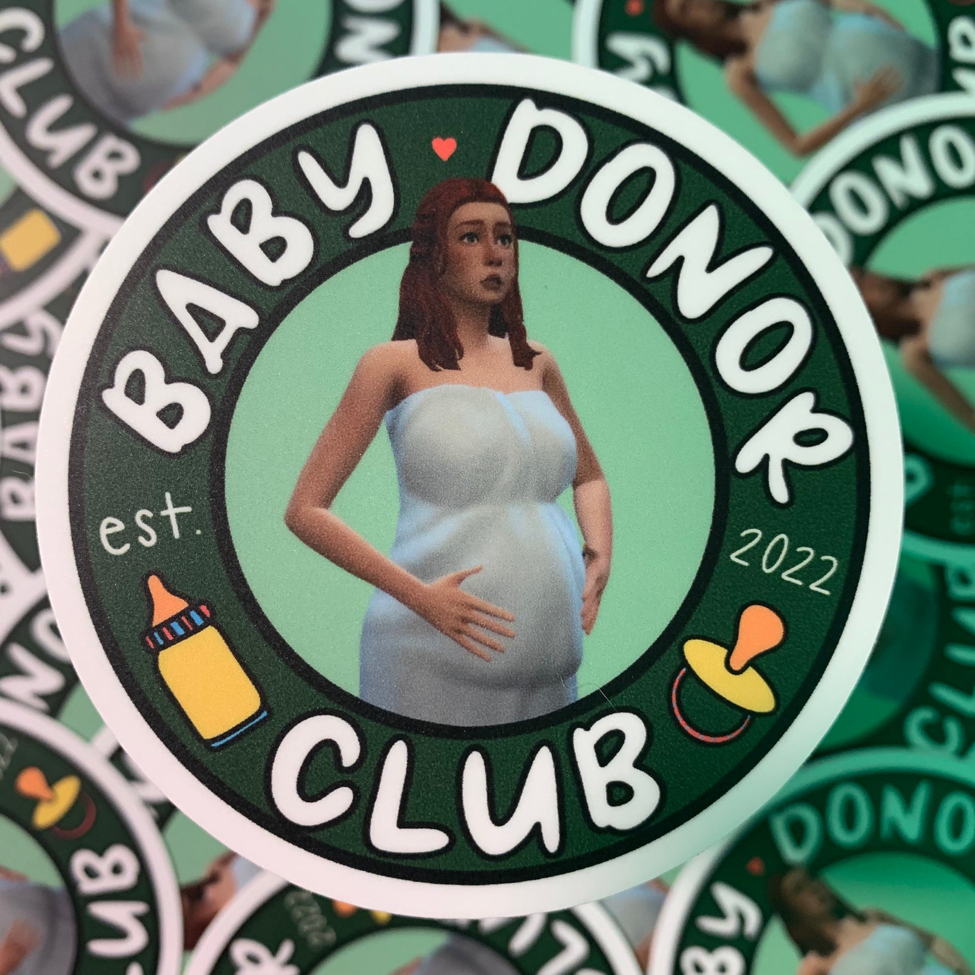 A circular sticker design with BABY DONOR CLUB written on it, with an image of "Hot Donna" in the middle. (Hot Donna is the matriarch from Bella's 100 baby challenge). There is also a small drawing of a baby bottle and a pacifier as well as est. 2022 on the design.