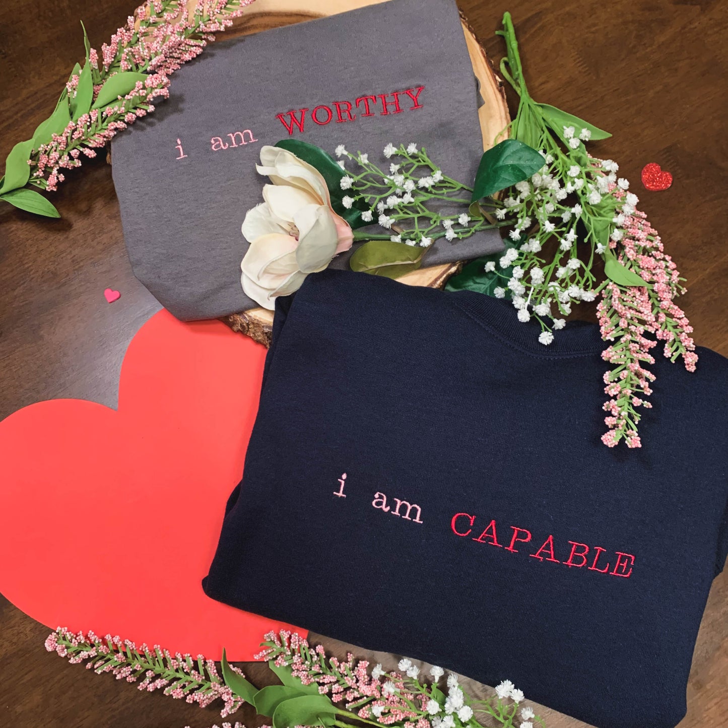 Overhead view of the affirmation clothing items, both folded with pink and white flowers around. The affirmation t-shirt is shown in gray with the embroidery "i am WORTHY" in pink and red. The affirmation crewneck sweatshirt is shown in navy blue with the embroidery "i am CAPABLE" in pink and red.