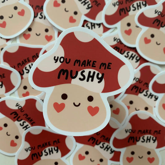 A sticker of a cartoon mushroom with a small smiley face, with heart shaped blush on the cheeks, with the phrase "you make me mushy" in all caps on the mushroom cap
