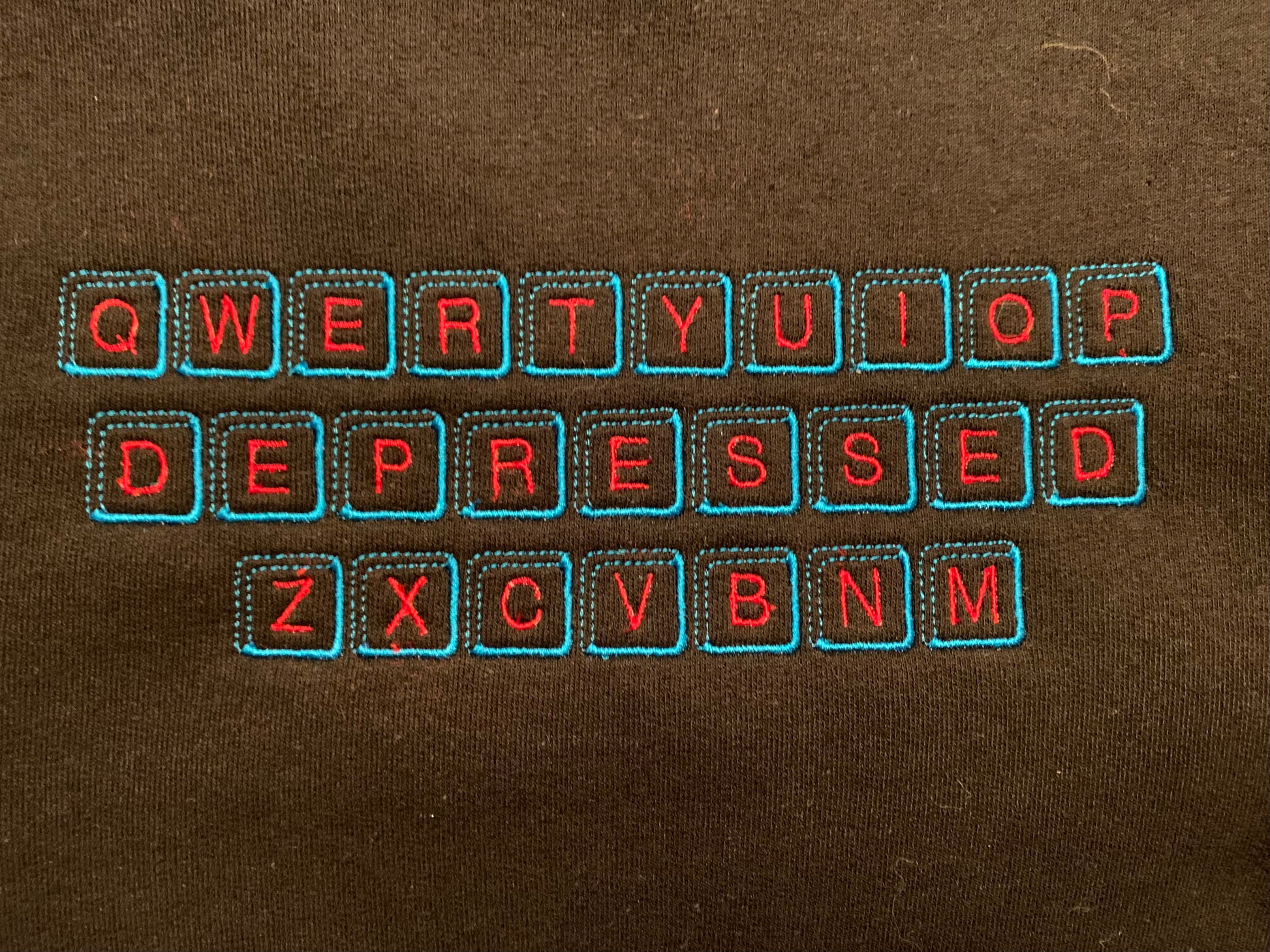 An upclose picture of a black crew neck with an embroidered design inspired by a qwerty keyboard on it. In the design, the middle row of keys are replaced with keys that spell DEPRESSED.  The key caps are embroidered in a light blue thread, with the letters on the caps embroidered in a bright red thread.