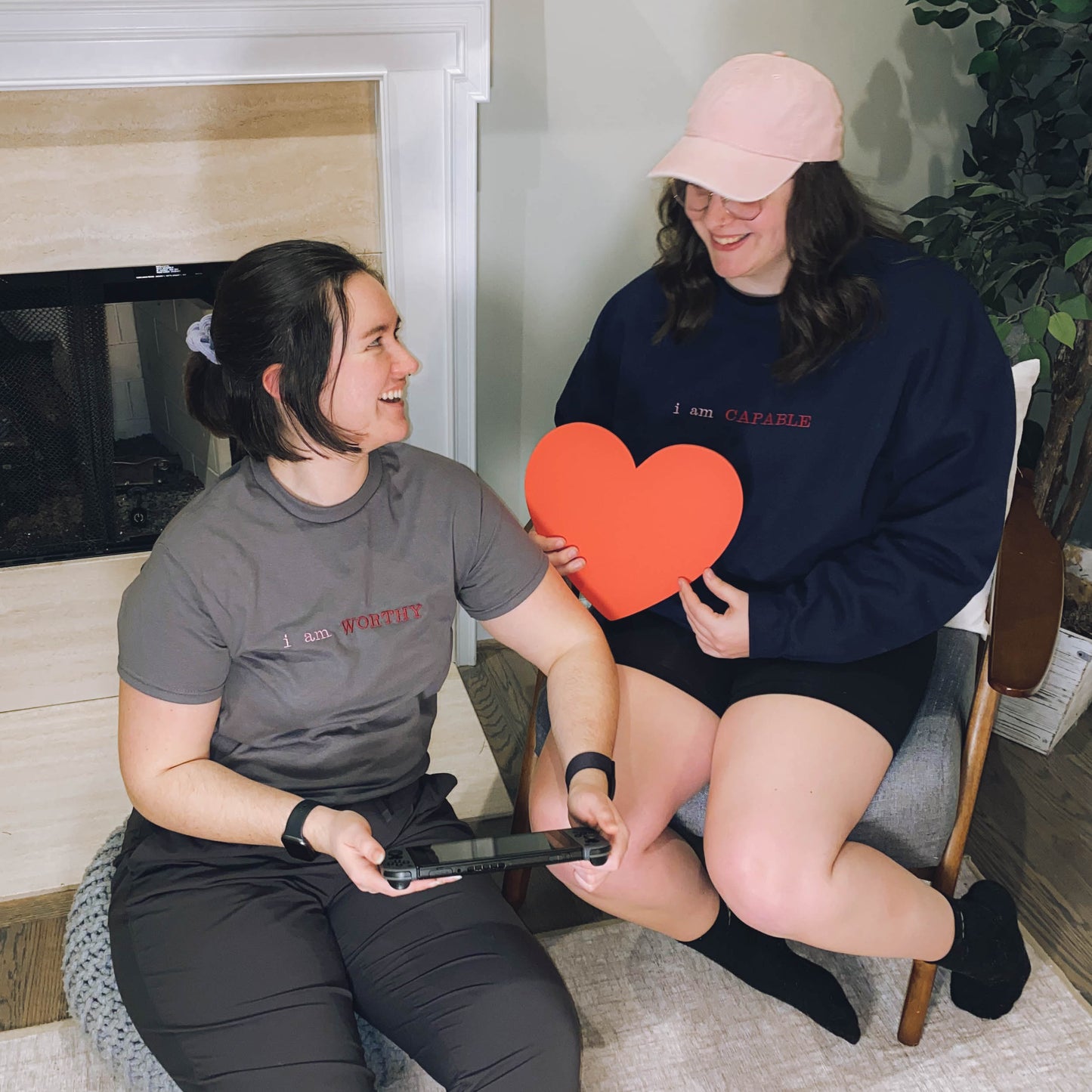 Bella and Ariel wearing the affirmation clothing items and smiling at each other. Bella is wearing the affirmation t-shirt in gray in size small with the embroidery "i am WORTHY" in pink and red. Ariel is wearing the affirmation crewneck sweatshirt in navy blue in size 2XL with the embroidery "i am CAPABLE" in pink and red.