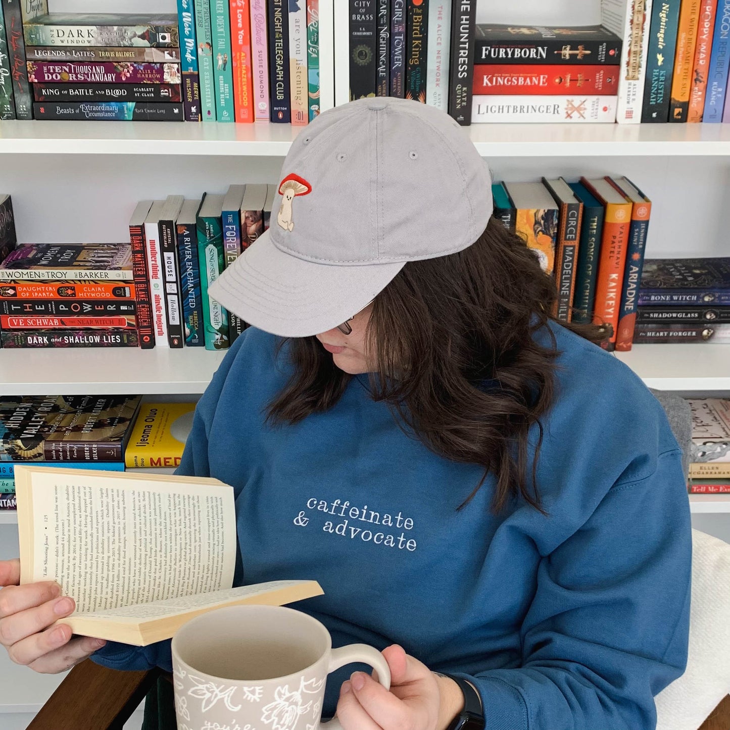 Ariel sitting in front of a bookcase reading and holding a coffee mug. She is wearing an indigo blue crew neck that is embroidered "caffeinate & advocate" on the front. Ariel is featured wearing a size Large.