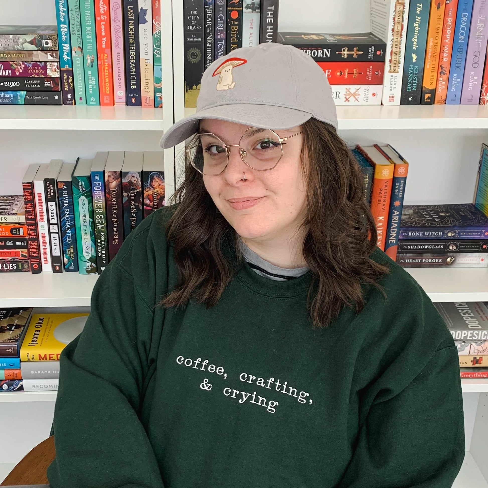 Ariel is sitting in front of a bookcase, looking at the camera and gently smiling. She is wearing a forest green crew neck that is embroidered "coffee, crafting, & crying" on the front. Ariel is featured wearing a size XL.