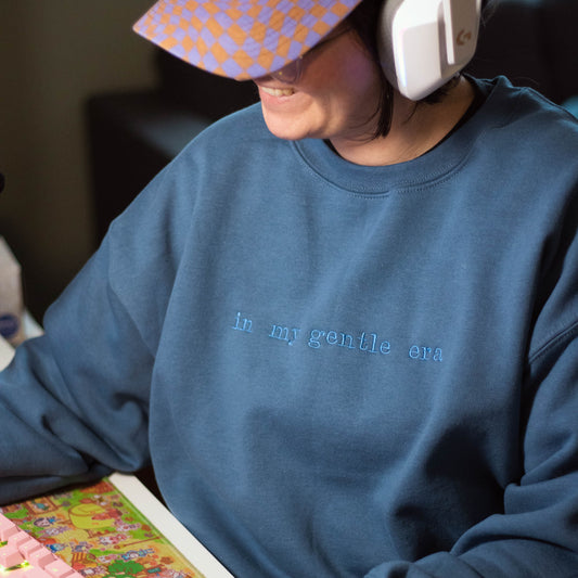 Bella sitting at a desk looking to the side wearing the blue gentle era crewneck sweatshirt. The sweatshirt is a slate blue color with slate blue embroider on the front that says "in my gentle era". Bella is wearing a size large.