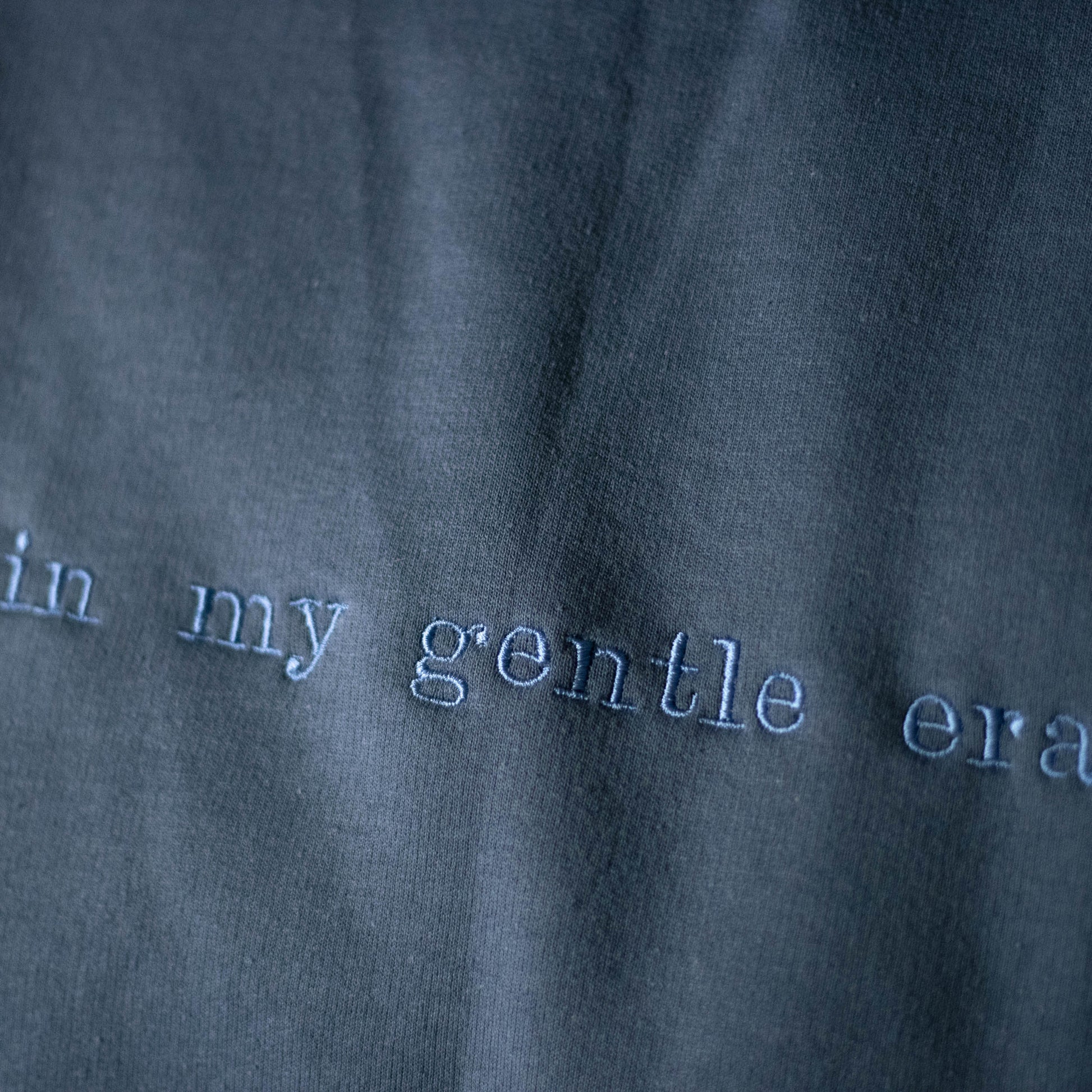 Close up image of the embroidered words on the blue gentle era crewneck sweatshirt. The sweatshirt is a slate blue color with slate blue embroidery on the front that says "in my gentle era".