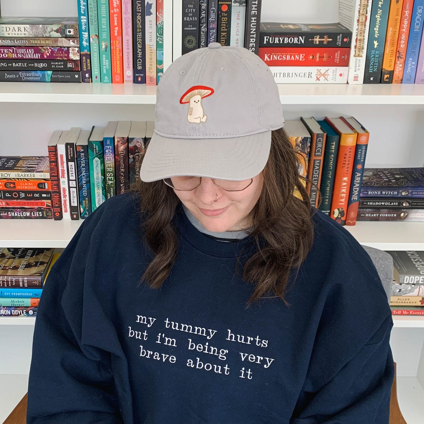 Ariel sitting in front of a bookcase looking down and smiling. She is wearing a navy blue crew neck that is embroidered "my tummy hurts but i'm being very brave about it" on the front. Ariel is featured wearing a size 2XL.