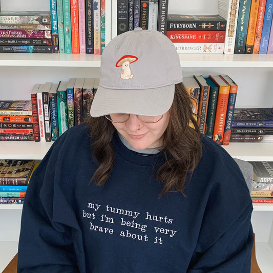 Ariel sitting in front of a bookcase looking down and smiling. She is wearing a navy blue crew neck that is embroidered "my tummy hurts but i'm being very brave about it" on the front. Ariel is featured wearing a size 2XL.