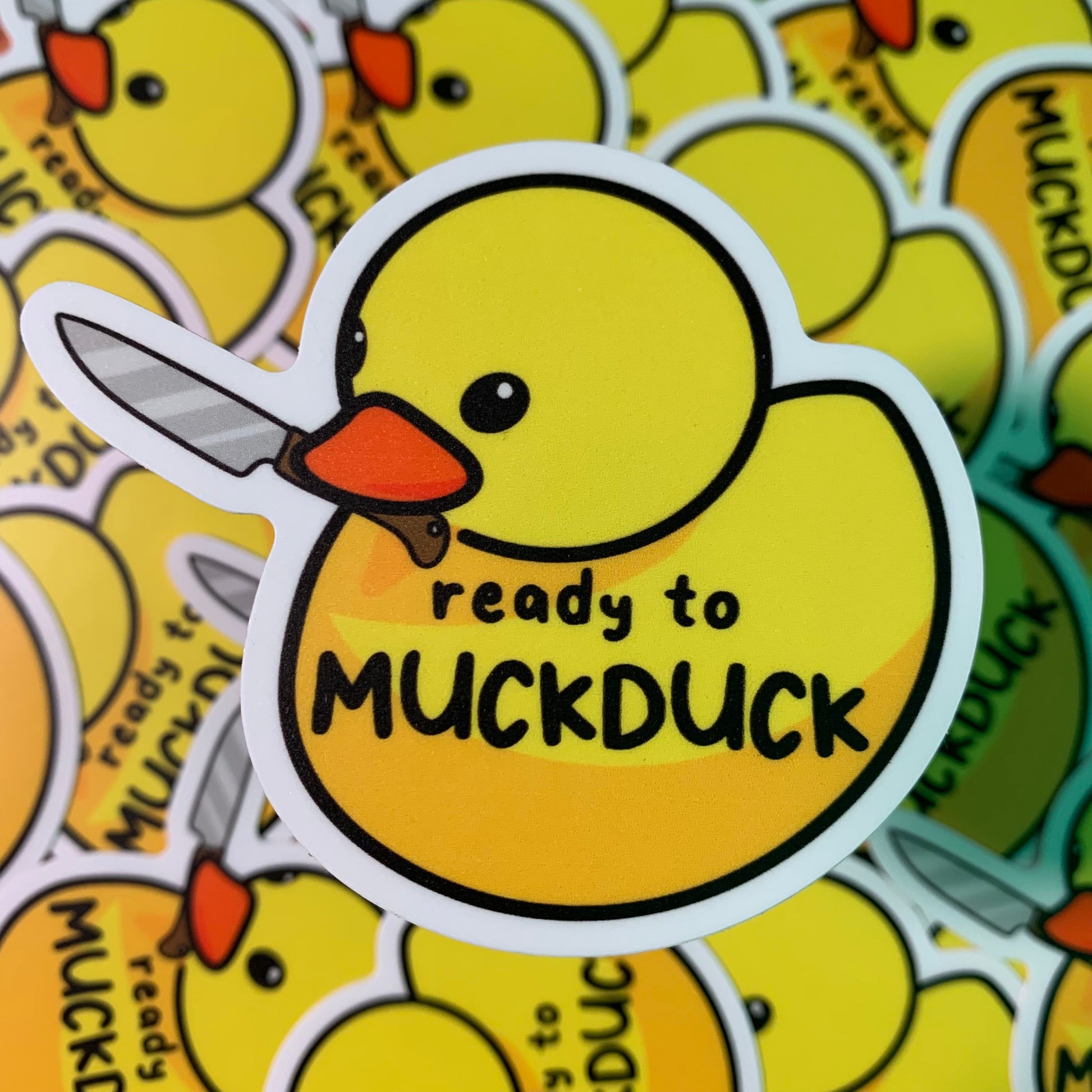 Sticker of a drawn plastic duck holding a kitchen knife, with the words "ready to MUCKDUCK" written on the front.